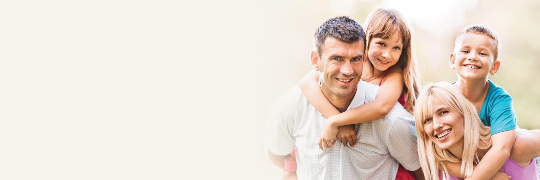 A happy family of four, including a father, mother, daughter, and son, hugging and smiling outdoors.